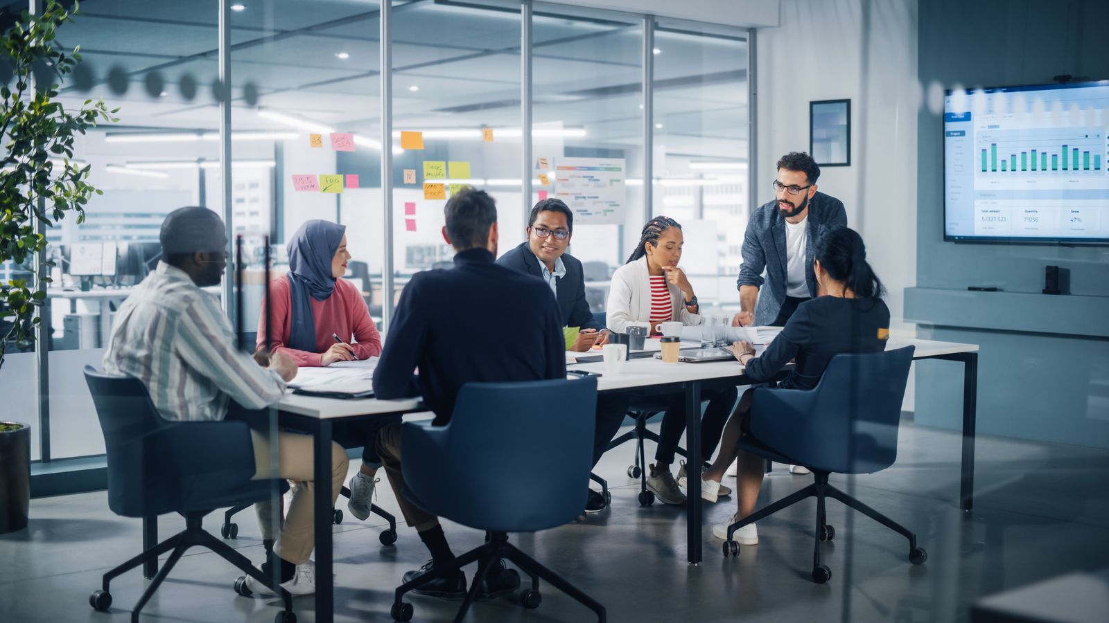 A team of colleagues in discussion around a meeting table in a glass-walled office, with one standing to lead the conversation